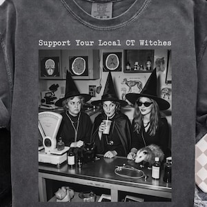 Könnte beinhalten: Ein anthrazitfarbenes T-Shirt mit einem schwarz-weißen Aufdruck von drei Personen mit Hexenhüten und dem Text "Support Your Local CT Witches". Die Grafik zeigt eine Laborszene mit einer Waage, einer Katze und einem Opossum auf einem Tisch.