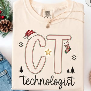 Könnte beinhalten: Ein elfenbeinfarbenes T-Shirt mit den Buchstaben CT in rot-grüner Umrandung, einer Weihnachtsmütze auf dem C, einem Weihnachtsstrumpf am T, einem goldenen Stern in der Mitte und dem Wort technologist darunter. Das Design zeigt kleine Schneeflocken.