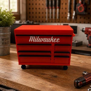 May include: A red Milwaukee tool chest with black drawer accents and the Milwaukee logo in white. The tool chest is on wheels and sits on a wooden surface. The background includes tools and a pegboard.