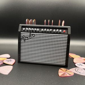 May include: A black miniature amplifier with the Fender logo, a silver speaker grill, and several knobs. Guitar picks in various colors and designs surround the amplifier, some with text. The amplifier has a red power indicator light.