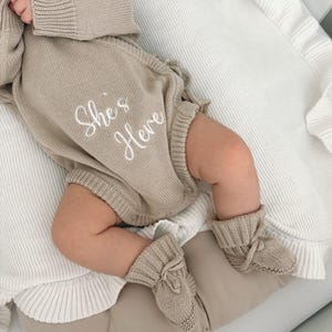 Peut inclure: Ensemble pour bébé en tricot beige comprenant un barboteuse et des chaussons assortis. La barboteuse porte l'inscription brodée "She's Here" en écriture blanche. L'ensemble est présenté sur une couverture blanche et beige.