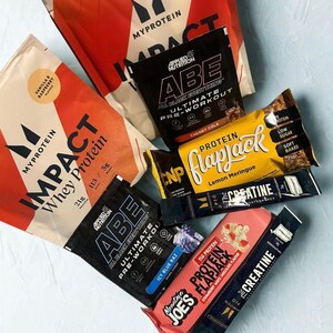 May include: An assortment of protein supplements and energy bars. The image features several packages of Myprotein Impact Whey Protein, Applied Nutrition ABE pre-workout, and CNP Protein Flapjack bars. Flavors include vanilla raspberry, cherry cola, and lemon meringue.