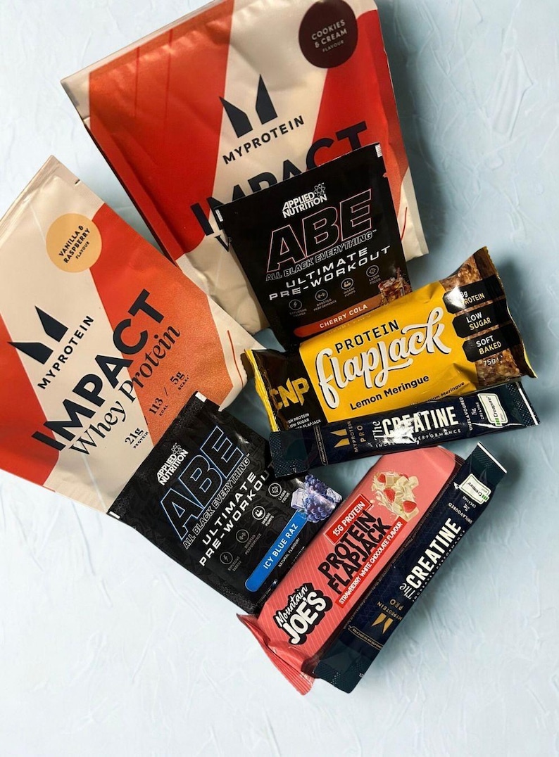 May include: An assortment of protein supplements and energy bars. The image features several packages of Myprotein Impact Whey Protein, Applied Nutrition ABE pre-workout, and CNP Protein Flapjack bars. Flavors include vanilla raspberry, cherry cola, and lemon meringue.