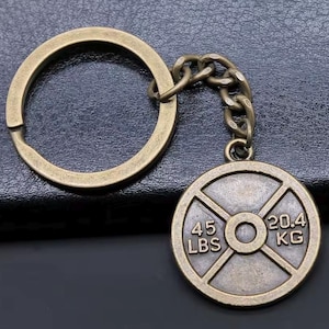 May include: Bronze-coloured weight plate keyring. The weight plate has the numbers "45 LBS" and "20.4 KG" printed on it. The keyring includes a chain and a large key ring.