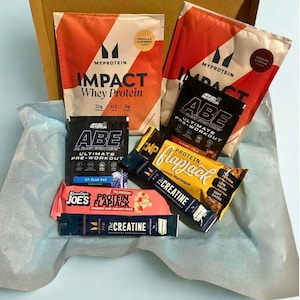 May include: A cardboard box filled with various protein supplements and snacks. The box contains two bags of Myprotein Impact Whey Protein, two Applied Nutrition ABE pre-workout sachets, two protein flapjack bars, and a creatine sachet. The products are arranged on blue tissue paper.