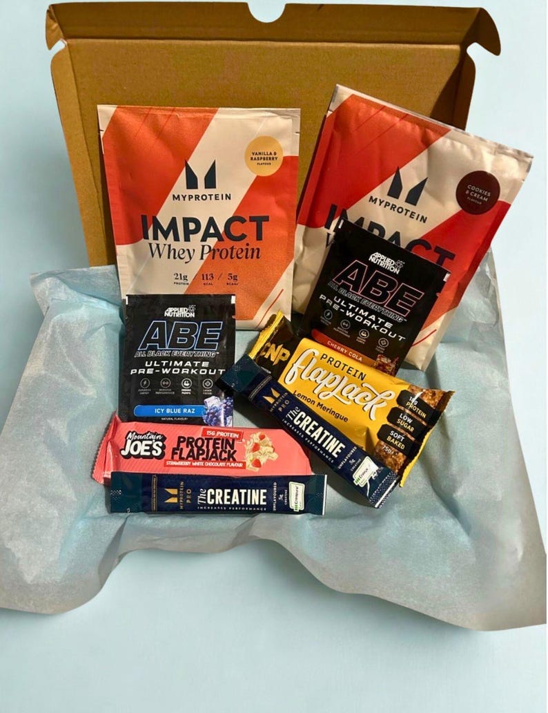 May include: A cardboard box filled with various protein supplements and snacks. The box contains two bags of Myprotein Impact Whey Protein, two Applied Nutrition ABE pre-workout sachets, two protein flapjack bars, and a creatine sachet. The products are arranged on blue tissue paper.