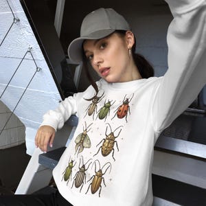 Puede incluir: Sudadera blanca con una ilustración de insectos en varios colores, incluyendo verde, marrón y rojo. El diseño incluye escarabajos y otros insectos. La sudadera de manga larga tiene cuello redondo.