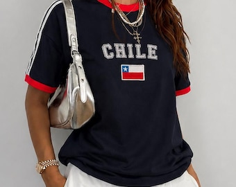 May include: A navy blue short-sleeved t-shirt with red trim on the sleeves and neckline, featuring the word CHILE in white block letters above a small embroidered Chilean flag. The shirt is paired with a metallic silver shoulder bag.