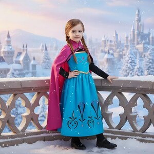 May include: A young girl in a blue dress with black sleeves and a pink cape, posing on a snowy balcony. The dress features a gold trim and decorative floral patterns. A castle is visible in the background.
