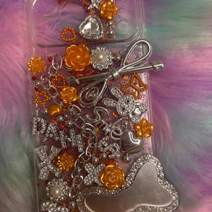 May include: Clear phone case decorated with silver and gold embellishments. The case features a variety of charms, including flowers, hearts, and a bow. The word "DAHLIA" is spelled out in silver letters. The background is a fluffy, multi-colored fabric.