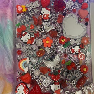 Könnte beinhalten: Eine transparente Handyhülle, verziert mit verschiedenen roten und silbernen Charms. Zu den Charms gehören Hello Kitty-Figuren, Erdbeeren, Herzen, Schleifen, Blumen und eine silberne Kette. Das Wort "Smile" ist sichtbar.