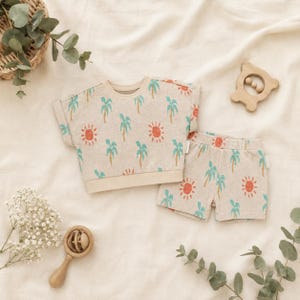 May include: A two-piece outfit consisting of a short-sleeved top and shorts in a light beige fabric. The set features a repeating pattern of teal palm trees and orange suns with simple facial features. The top has a crew neckline and ribbed hem.