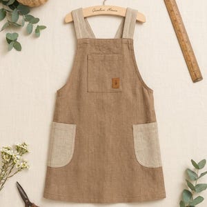 Puede incluir: Un delantal marrón con tirantes de color beige claro y dos bolsillos laterales a juego. El delantal cuenta con un bolsillo en el pecho con un pequeño parche rectangular marrón que muestra un icono de árbol. Cuelga de una percha de madera con la etiqueta Gumbui House.