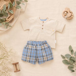 May include: A beige short-sleeved henley shirt with three buttons and a chest pocket, paired with blue and tan plaid shorts featuring an elastic waistband and white drawstring. The set is displayed alongside a wooden rattle and eucalyptus leaves.