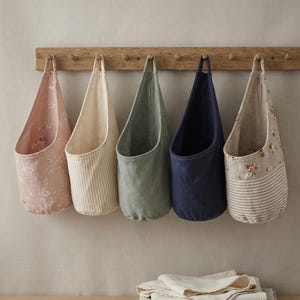 May include: Five fabric hanging storage baskets suspended from a wooden peg rail. The baskets feature various designs including a pink floral print, beige vertical stripes, solid sage green, solid navy blue, and a combination of floral and striped patterns.