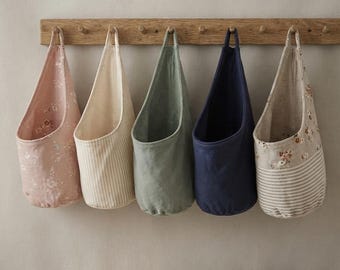 Può includere: Cinque cesti portaoggetti in tessuto appesi a un binario con pioli in legno. I cesti presentano vari design, tra cui una stampa floreale rosa, strisce verticali beige, verde salvia tinta unita, blu navy tinta unita e una combinazione di motivi.
