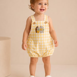 May include: A yellow and white gingham romper featuring two wooden buttons at the shoulder straps and an embroidered graphic on the chest depicting a small hedgehog and a bear holding green sprigs, paired with white strappy sandals.