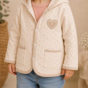 Puede incluir: Una chaqueta con capucha de color crema con un delicado estampado floral rosa y verde, un parche en forma de corazón en el pecho con flores bordadas, dos bolsillos delanteros, ribete beige y botones a presión blancos, sobre una camisa rosa.