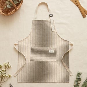 May include: A kitchen apron featuring a vertical dark grey and off-white pinstripe pattern. The apron includes a single front pocket with a small rectangular white label, an adjustable neck strap with a metal slider, and long side ties.