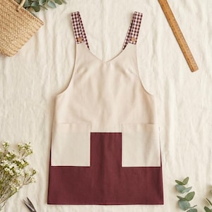 Può includere: Un grembiule bicolore con una sezione superiore color crema e una sezione inferiore bordeaux scuro. Il grembiule presenta due tasche frontali e spallacci con motivo a quadretti marrone e bianco, fissati da piccoli bottoni. È visibile un righello in legno.