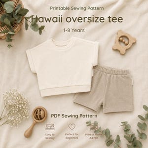 May include: A printable sewing pattern for a Hawaii oversize tee for ages 1-8 years. The image displays a cream-colored short-sleeved shirt and beige shorts, accompanied by a wooden rattle and a wooden teether on a neutral fabric background.