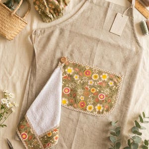 May include: A beige linen apron featuring a front pocket with a green floral pattern, a matching detachable white terry cloth towel secured by a wooden button, and a coordinating green floral hair bonnet, all displayed with sewing accessories.