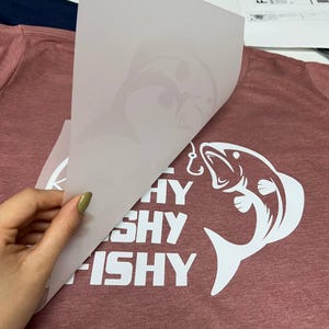 Puede incluir: Una camiseta granate con un gr&aacute;fico blanco de un pez y las palabras "SHY FISHY FISHY". Una mano despega una l&aacute;mina de transferencia blanca, revelando el dise&ntilde;o. La camiseta est&aacute; hecha de un material suave y texturizado.