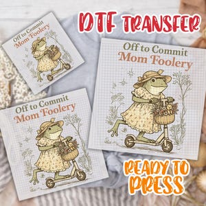 May include: DTF transfer featuring a whimsical frog riding a scooter, wearing a dress and hat, with a basket of flowers. The text "Off to Commit Mom Foolery" is displayed on a grid background. The words "Ready to Press" are also visible.