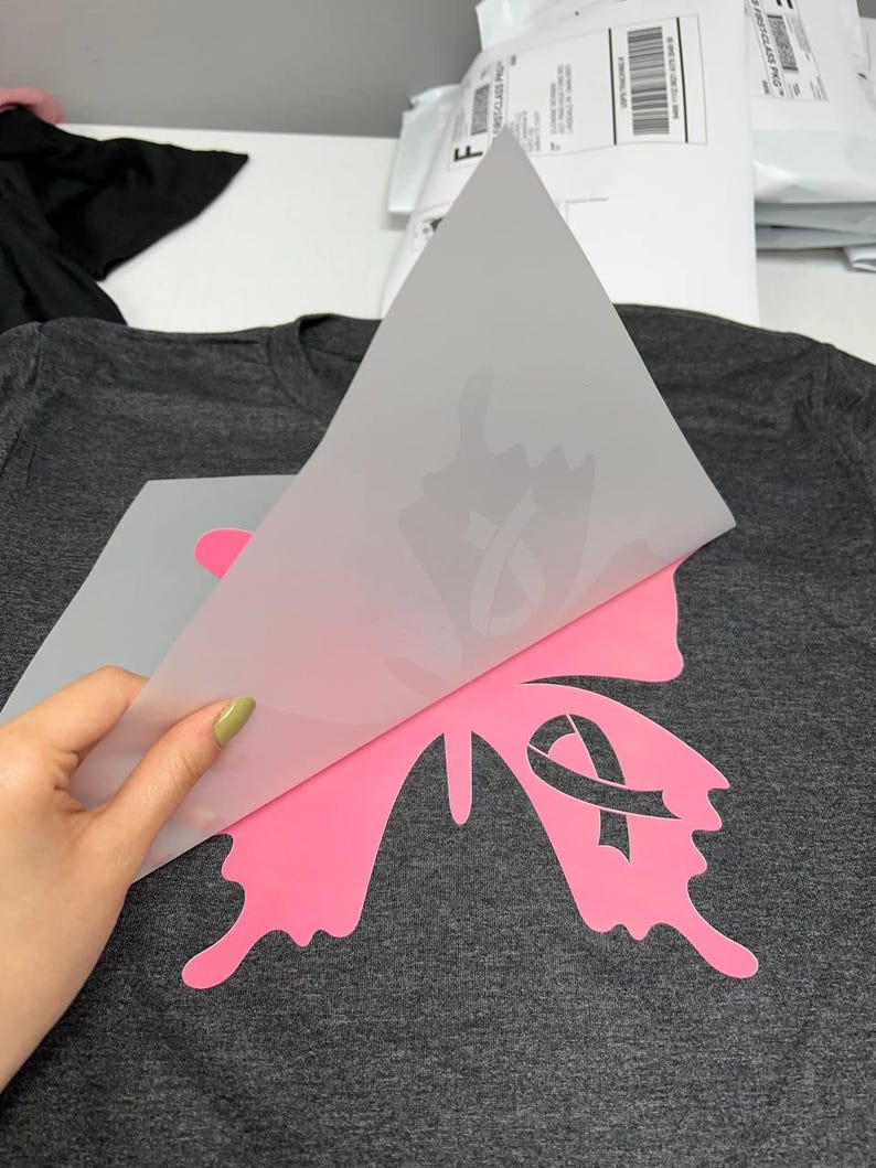 Puede incluir: Una camiseta gris oscuro con un dise&ntilde;o de mariposa rosa con una cinta rosa. El dise&ntilde;o se aplica a la camiseta mediante transferencia t&eacute;rmica. El dise&ntilde;o de la mariposa es un s&iacute;mbolo de concienciaci&oacute;n sobre el c&aacute;ncer de mama.