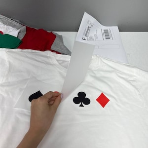 Puede incluir: Camiseta blanca con s&iacute;mbolos negros de tr&eacute;bol y pica, y un s&iacute;mbolo de diamante rojo. Una mano levanta una hoja de transferencia. Otras camisetas son visibles en el fondo.