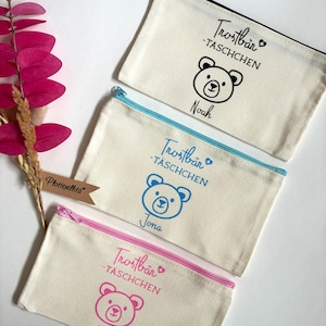 Puede incluir: Tres estuches con cremallera de lona con diseños de osos y nombres. El estuche superior tiene una cremallera negra y un gráfico de oso negro con el nombre "Noah". El estuche del medio tiene una cremallera azul y un gráfico de oso azul con el nombre "Jona". El estuche inferior tiene una cremallera rosa y un gráfico de oso rosa con el nombre "Mila".