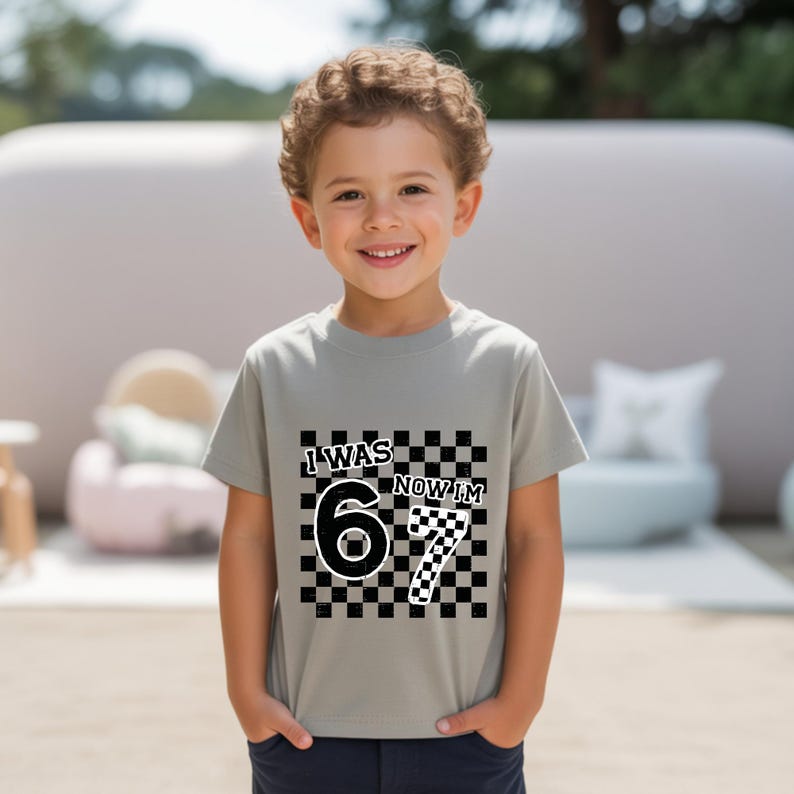 May include: A gray t-shirt with the text "I WAS 6 NOW I'M 7" in black and white. The number 6 is solid black, and the number 7 is a black and white checkered pattern. The shirt is for a child.