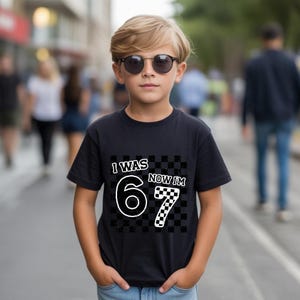 May include: A black t-shirt with the text "I WAS 6 NOW I'M 7" in white and checkered patterns. The shirt is worn by a young person wearing sunglasses, standing on a city street.