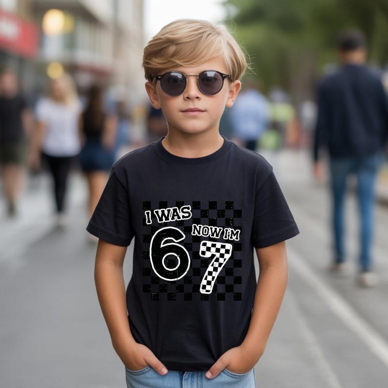 May include: A black t-shirt with the text "I WAS 6 NOW I'M 7" in white and checkered patterns. The shirt is worn by a young person wearing sunglasses, standing on a city street.