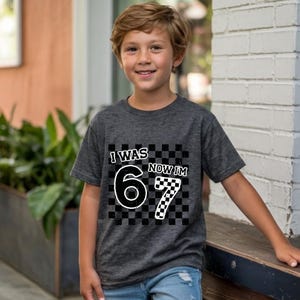 May include: Dark gray t-shirt with the text "I WAS 6 NOW I'M 7" in a black and white checkered design. The shirt is short-sleeved and made of a soft material. The shirt is suitable for a child's birthday.