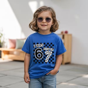 May include: A blue t-shirt with the text "I WAS 6 NOW IM 7" in a black and white checkered design. The shirt is worn by a child wearing sunglasses.
