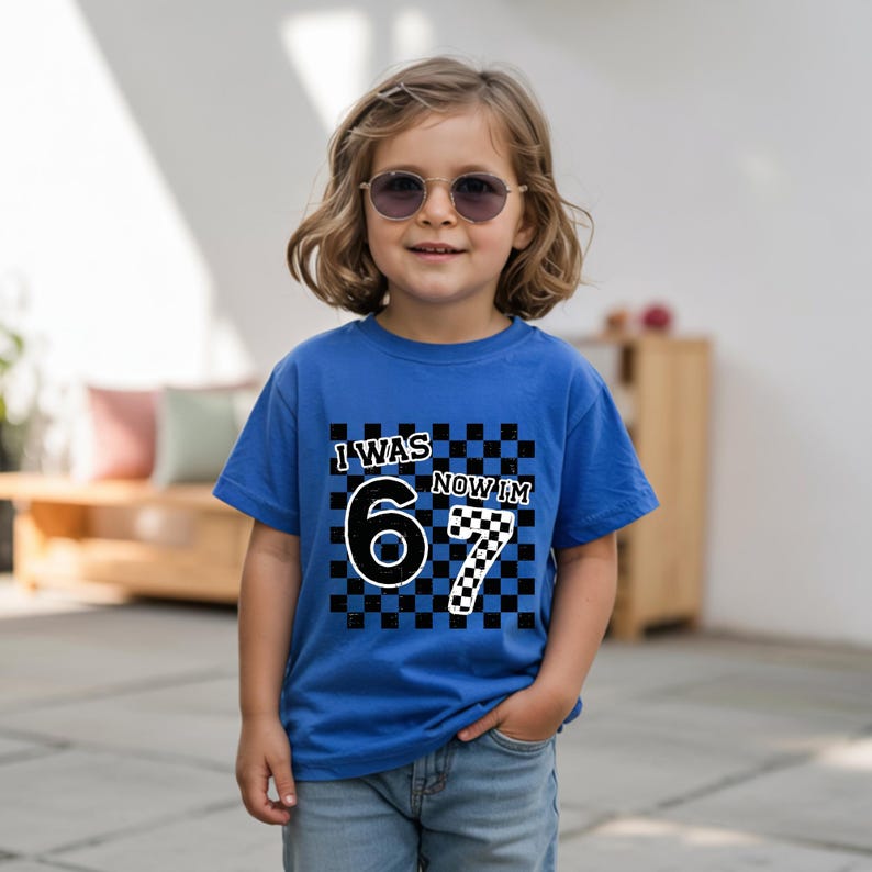 May include: A blue t-shirt with the text "I WAS 6 NOW IM 7" in a black and white checkered design. The shirt is worn by a child wearing sunglasses.