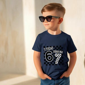 May include: Navy blue t-shirt with the text "I WAS 6 NOW I'M 7" in a black and white checkered design. The shirt is worn by a young child wearing black sunglasses.