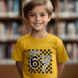 May include: A yellow t-shirt with the text "I WAS 6 NOW I'M 7" in black and white. The number 6 is in a large, bold font, and the number 7 is in a checkered pattern. The shirt is worn by a smiling child.