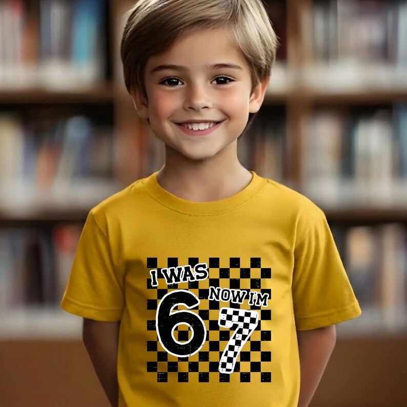 May include: A yellow t-shirt with the text "I WAS 6 NOW I'M 7" in black and white. The number 6 is in a large, bold font, and the number 7 is in a checkered pattern. The shirt is worn by a smiling child.