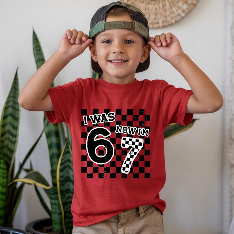 May include: Red t-shirt with the text "I WAS 6 NOW I'M 7" in a black and white checkered design. The shirt is worn by a child wearing a camouflage baseball cap.