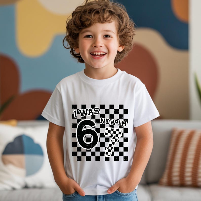 May include: A white t-shirt with the text "I WAS 6 NOW I'M 7" in a black and white checkered design. The shirt is worn by a smiling child. The shirt is a birthday-themed apparel.