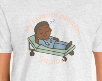 May include: A light gray t-shirt featuring a cartoon illustration of a person in blue scrubs lying on a medical bed. The text on the shirt reads "My favorite position Supine". The design includes a stethoscope.