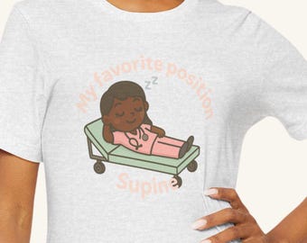 Pode incluir: T-shirt cinza claro com uma ilustração de desenho animado de uma enfermeira dormindo em uma cama médica. A enfermeira está usando um uniforme rosa e um estetoscópio. O texto "My favorite position" e "Supine" está impresso na camiseta.