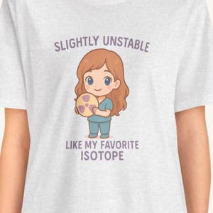 May include: White t-shirt with a cartoon graphic. The graphic features a smiling character holding a radiation symbol, with the text "SLIGHTLY UNSTABLE LIKE MY FAVORITE ISOTOPE" in purple and white.