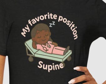 Pode incluir: Camiseta preta com uma ilustração de desenho animado de uma pessoa de uniforme rosa deitada em uma cama médica. O texto "My favorite position" está acima, com "Supine" abaixo.