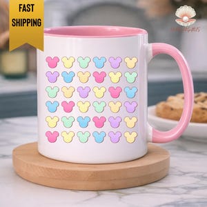 A white mug with a pink handle, featuring a repeating pattern of colorful Mickey Mouse-shaped icons on its surface.