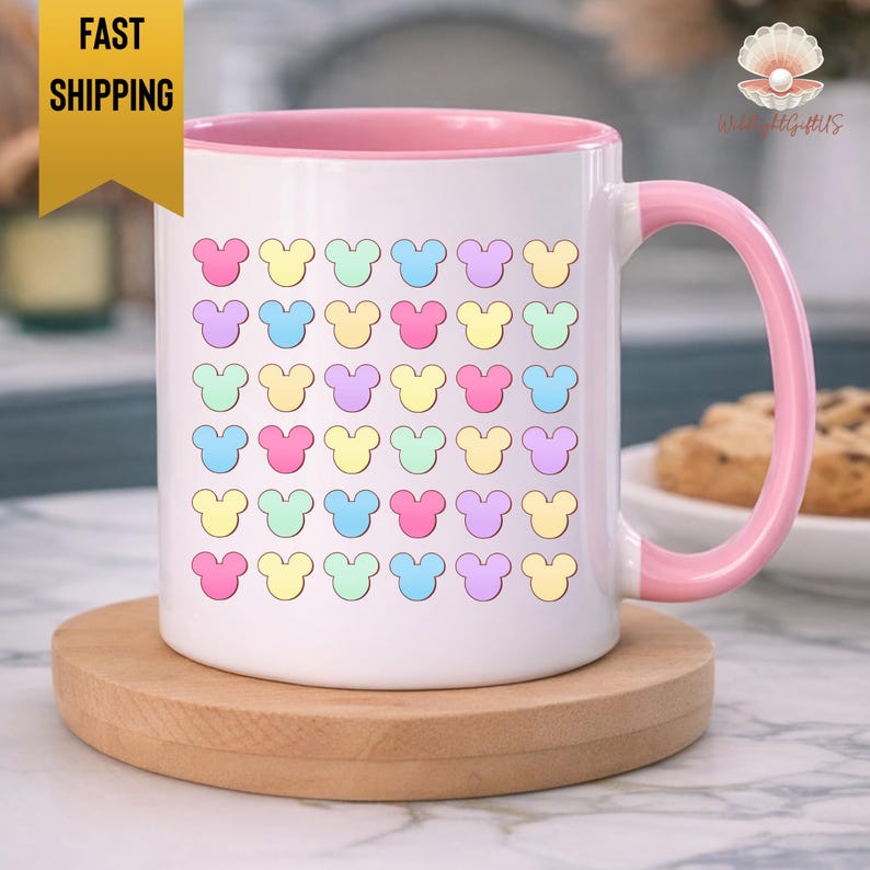 A white mug with a pink handle, featuring a repeating pattern of colorful Mickey Mouse-shaped icons on its surface.