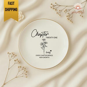 May include: A white ceramic plate with a gold rim, featuring the text "Chapter Twenty-One" and a floral illustration. The plate also includes the name "Emily" and the phrase "Every chapter brings new growth."