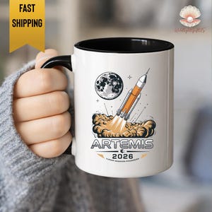 May include: A white ceramic mug with a black interior and handle. The mug features a graphic of a rocket launching towards the moon, with the text "ARTEMIS 2026 LUNAR MISSION PROGRESS".